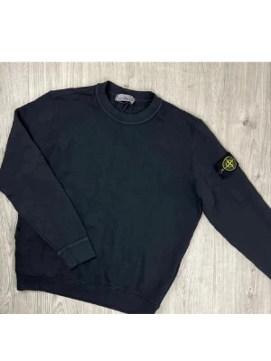 Svart sweatshirt från Stone Island - Svart långärmad sweatshirt från Stone Island med klassisk rund hals och ribbade muddar. Ikonisk Stone Island patch med broderad logga i gult och grönt sitter på vänster ärm. Perfekt för dig som gillar streetwear och vill ha en clean look.
