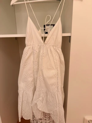 Vit somrig klänning med spetsdetaljer - Säljer en vit kortklänning med tunna axelband och v-ringad front. Klänningen har broderade spetsdetaljer och volang nedtill, vilket ger en romantisk och fräsch känsla. Perfekt för varma dagar och har en lätt, luftig passform.