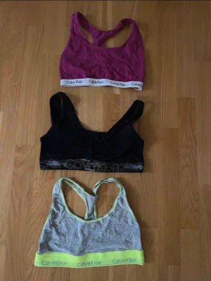 3 Calvin Klein bh-toppar - Tre snygga bh-toppar från Calvin Klein i olika färger: en lila med vit logokant, en svart med diskret logga och en grå med limegrön kant och logga. Alla har racerback och är perfekta för träning eller chill. Mjukt bomullsmaterial och sportig design.