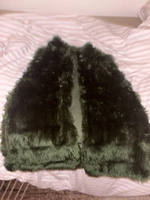 Mörkgrön fluffig pälsväst - Säljer en riktigt mjuk och fluffig mörkgrön pälsväst. Västen har en rak och klassisk form utan krage eller synliga detaljer, och är helfärgad i en djup grön ton. Perfekt att slänga över en hoodie eller långärmad tröja för extra värme.