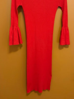 Röd ribbad klänning med volangärm - Säljer en snygg röd ribbad klänning med långärmade ärmar som avslutas med breda volanger. Klänningen har en kroppsnära passform och är tillverkad i ett stickat material som känns mjukt och följsamt. Perfekt för dig som gillar färg och coola detaljer.