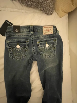 Oanvända True Religion blå bootcut jeans W26 - HELT NYA snygga blå jeans från True Religion med bootcut passform och klassiska kontrastsömmar. Modellen heter Becca och har detaljerade fickor med lock och silverfärgade knappar. Jeansen har låg midja och är tillverkade i slitstarkt denim med gul söm.