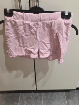 Ljusrosa shorts från ONLY - Säljer ett par ljusrosa shorts från ONLY med elastisk midja och trycket 'HAWAII' samt broderade blommor på ena benet. Sköna och mjuka, perfekta för varma dagar. Materialet är bomull och modellen är loose fit.