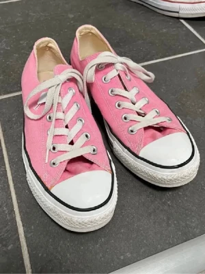 Rosa Converse sneakers med vita snören - Säljer ett par klassiska Converse sneakers i rosa canvas med vita snören och tåhätta i gummi. Skorna har låg profil, rund tå och vit sula med svart rand. Perfekta för dig som vill ha en färgklick till vardagslooken.