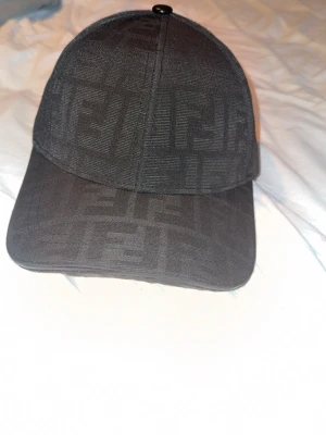 Svart Fendi keps med mönster - Snygg svart keps från Fendi med diskret mönster över hela ytan och justerbar rem baktill. Insidan har Fendi-loggor på banden. Perfekt för dig som vill ha en stilren accessoar med lyxig känsla.