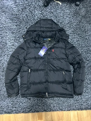 Svart pufferjacka från Polo Ralph Lauren - Svart pufferjacka från Polo Ralph Lauren med huva och två sidofickor med dragkedja. Jackan har det klassiska röda Polo-logot broderat på bröstet och quiltad design. Perfekt för kalla dagar och har en snygg, sportig vibe.