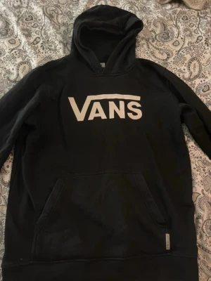 Vans hoodie (liten i storleken, sliten) - Denna Vans hoodie är märkt som storlek XL, men den är liten i storleken och passar mer som en small eller medium. Den är lite sliten men inget större problem, den ger en cool vintage känsla. Perfekt för en avslappnad stil eller att mysa runt hemma. Färgen är svart och den har det klassiska Vans-logotypen på framsidan.