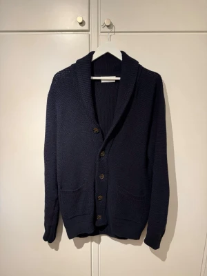 Selected Homme Marinblå Knappkofta – Klassisk Smart Casual Stickning - Klassisk kofta med knappar från Selected Homme i en tidlös marinblå färg. Ett mångsidigt plagg som fungerar perfekt för både smart casual och vardagsbruk.  Storlek L Bredd: 52 cm Längd: 73 cm Ärm från axel: 72 cm  Stickningen har en strukturerad men ändå bekväm känsla, med knappar fram, två framfickor och en ren sjalkrage. Lätt att styla över en skjorta, t-shirt eller stickad klänning för en elegant skandinavisk look.  Liten (!) defekt på baksidan (se bild 4) Annars är den endast använd 2 gånger