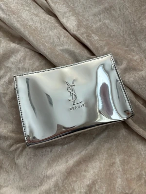 Silver sminkväska från YSL Beauté - Snygg och glansig sminkväska från YSL Beauté i metallic silver. Väskan har en stilren, rektangulär form och är tillverkad i syntetmaterial med YSL-logga i mitten. Perfekt för dig som vill ha en statement-accessoar med lyxig känsla.
