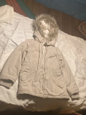 Beige parkajacka med fuskpäls från H&M - Snygg beige parkajacka från H&M L.O.G.G. med fluffigt fuskpälsfoder i luvan, dragkedja och snörning i midjan. Jackan har två fickor med knappar och ribbade muddar vid ärmsluten. Perfekt för kalla dagar och chill stil.
