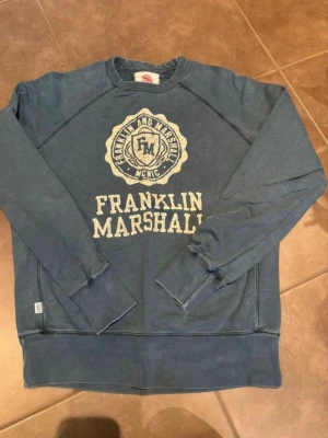 Blå sweatshirt från Franklin & Marshall - Säljer en blå sweatshirt från Franklin & Marshall i storlek XL. Tröjan har tryckt logga och text framtill, rund halsringning och långa ärmar. Tillverkad i mjuk bomull, perfekt för chill dagar eller när du vill ha en avslappnad stil.