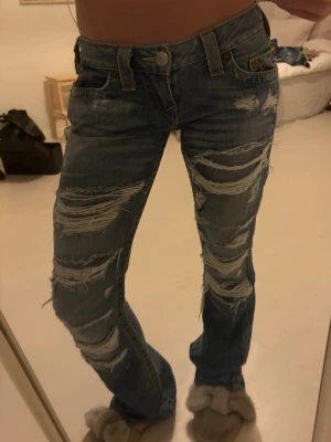 True Religion slitna bootcut jeans - Säljer ett par blå jeans från True Religion med bootcut passform och rejält slitna detaljer framtill. Klassisk femficksmodell med lågmidja. Det är storlek W25 men skulle säga att dom är större i storlek💕 skriv om ni har några funderingar💕