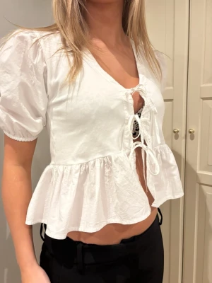 Vit blus med knyt och volang - Supersöt vit blus med puffärmar, v-ringning och två knyt framtill. Toppen har volang nedtill och är croppad.Tillverkad i ett lätt bomullstyg som känns luftigt.🤍
