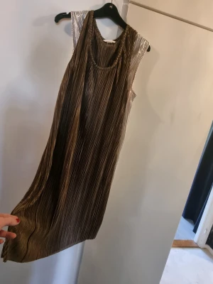 Plisserad metallic klänning - Säljer en plisserad klänning i metallicton med snyggt fall och ärmlös design. Klänningen har rundad halsringning och skimrar i guld och silver, perfekt för dig som vill sticka ut. Materialet känns lätt och glansigt.