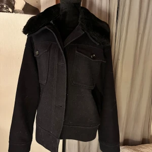Svart ulljacka med fuskpäls från Noella - Svart jacka från Noella i ullmaterial med fuskpäls på kragen. Jackan har två stora bröstfickor med knappar och en klassisk, lite loose passform. Perfekt för dig som gillar stilrena och varma jackor med coola detaljer.