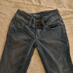 Blå bootcut jeans från Orsay - Säljer ett par blå jeans från Orsay med bootcut-snitt och låg midja. Jeansen har klassisk femficksdesign, kontrastsömmar och tre knappar i midjan. Materialet är jeans med en mjuk känsla och snygg tvätt. Perfekt för dig som gillar retrovibbar och stilren look.