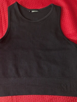 Svart ribbad linnetopp från Stronger - Svart croppad linnetopp från Stronger med ribbad struktur och bred kant nedtill där loggan är diskret broderad. Toppen är ärmlös och har rund halsringning, perfekt för träning eller chill. Materialet känns mjukt och stretchigt mot huden.