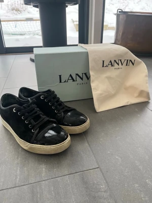 Svarta Lanvins - Lanvin sneakers i svart med snörning och rund tå. Ovandelen är i mocka och tåpartiet har en blank finish i skinn. Sulan är vit och platt. Medföljer originalkartong och dustbag från Lanvin. Köpt helt nya men använt ett tag. Som man kan se på bild 3 har det gått upp en bit på toecapen, men det syns ändast på nära håll. Den har gått upp lite i bakre delen av skon, men inget som märks vid användning. Skorna har mycket kvar att ge och är riktigt stilrena. Storlek 42, men sitter 43 Skriv vid intresse.