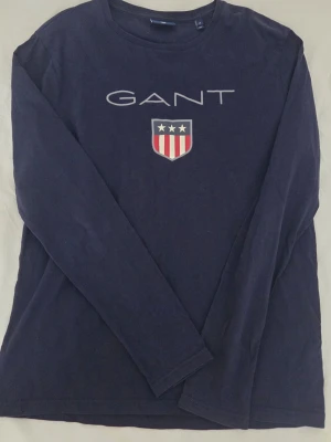 Marinblå långärmad topp från GANT - Säljer en marinblå långärmad topp från GANT i bomull. Toppen har rund hals och ett stort GANT-tryck med emblem på bröstet. Enkel och stilren design som funkar till jeans eller chinos. Perfekt för dig som gillar klassiska märken.