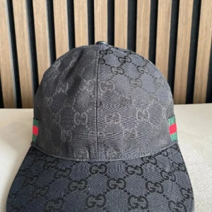Svart keps från Gucci med GG-mönster - Snygg svart keps från Gucci med klassiskt GG-monogram över hela kepsen. Kepsen har gröna och röda ränder på sidorna och justerbar rem baktill. Tillverkad i bomull och polyester, perfekt för dig som vill ha en exklusiv streetstyle-look.