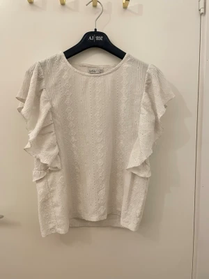 Vit blus med volangärm från Isolde - Säljer en söt vit blus från Isolde i storlek M. Blusen har fina volangärmar och ett diskret broderat mönster med små hål och geometriska detaljer. Materialet känns luftigt och perfekt för varmare dagar. Rund halsringning och normal passform.