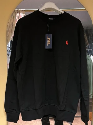 Svart sweatshirt från Polo Ralph Lauren - Svart sweatshirt från Polo Ralph Lauren med klassisk rund hals och långärmad design. Tröjan har en röd broderad logga på bröstet och är tillverkad i mjuk bomull. Perfekt för en chill och stilren look.