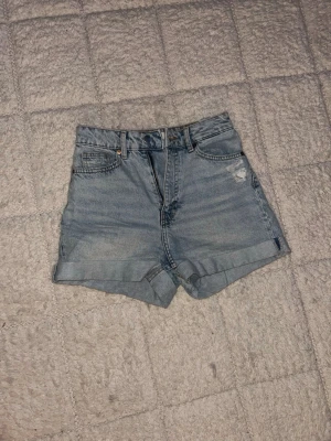 Ljusblå jeansshorts - Säljer ett par jeansshorts i ljusblå denim från H&M. Shortsen har en liten sliten detalj vid ena fickan och stängs med knapp och dragkedja. Perfekta för varma dagar och chill stil.