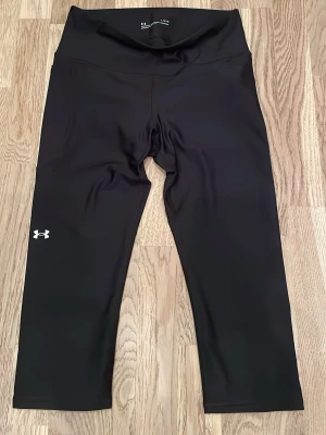 Svarta träningsleggings från Under Armour - Snygga svarta träningsleggings från Under Armour med klassisk logga på vänster ben. De är tillverkade i ett stretchigt syntetmaterial som sitter skönt och följsamt vid träning. Använda och tvättade en gång så nästan som nya. 