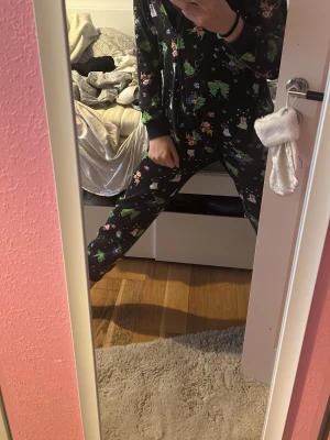 Mysiga julmönstrade byxdress i fleece - Säljer en svart byxdress i fleece med juligt mönster av granar, snögubbar och pepparkakor. Dragkedja framtill och en silverfärgad stjärna som detalj på luvan. Perfekt för chill dagar hemma under vintern.