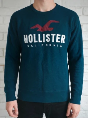 Hollister tröja  - Snygg mörkblå tröja från Hollister med stor vit logga och röd fågel på bröstet. Klassisk rund halsringning och långa ärmar. Tillverkad i mjukt material som passar perfekt till vardagslooken.