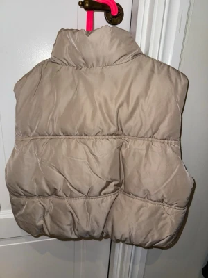 Beige väst med dragkedja - Säljer en beige väst från H&M med hög krage och dragkedja framtill. Västen har två fickor på sidorna och en puffig, quiltad design som ger en trendig look. Perfekt att slänga över en hoodie eller tröja för extra värme. 