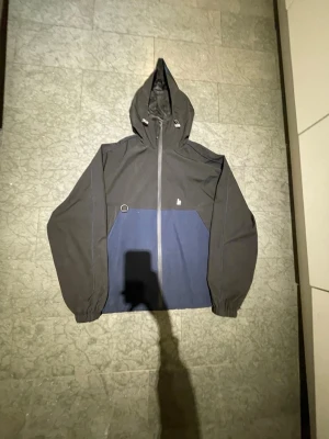 Harlaut jacka 1999 årsmodell 2023 - Säljer en stilren vindjacka från Harlaut Apparel Co i svart och marinblått. Jackan har huva med hel dragkedja framtill och broderad logga på bröstet samt tryck på ryggen. Materialet är och passformen är loose.