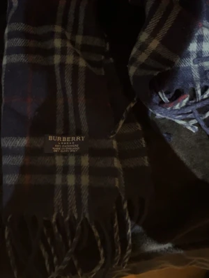 Rutig blå halsduk från Burberry - Säljer en klassisk rutig halsduk från Burberry i mörkblått, vitt och röda detaljer. Halsduken är tillverkad i 100% kashmir och har fransar i ändarna. Perfekt accessoar för att lyfta din outfit med en tidlös touch.