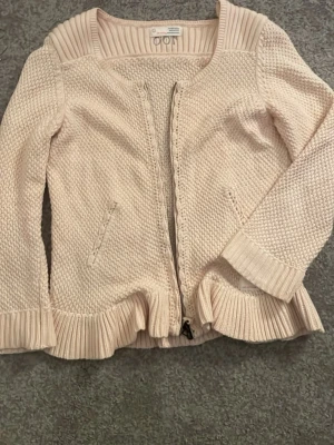 Ljusbeige kofta från Odd Molly - Supersöt ljusbeige kofta från Odd Molly med dragkedja framtill och volangkant nedtill. Stickad struktur och ribbade detaljer vid krage, ärmslut och nederkant. Perfekt för lager på lager och ger en chill men ändå söt vibe.