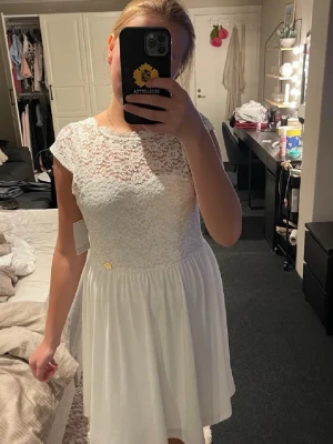 Vit spetsklänning med v-ringning bak - Superfin vit kortklänning med spets upptill och v-ringning i ryggen. Klänningen har korta ärmar och ett flowy kjolparti i mjukt tyg. Perfekt för sommarens alla tillfällen och har en liten guldfärgad detalj i midjan. Spetsen har blommigt mönster och ger en romantisk känsla.