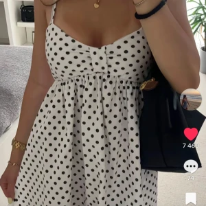 Vit prickig kort sommarklänning - Supersöt vit kortklänning med svarta prickar och smala axelband. Klänningen har en söt knutdetalj framtill och en lätt utställd kjol som ger en luftig känsla. Perfekt för varma dagar och ger en fräsch och lekfull look.