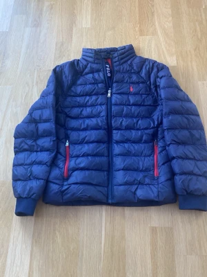 Blå pufferjacka från Polo Ralph Lauren - Snygg blå pufferjacka från Polo Ralph Lauren med röda detaljer och dragkedja framtill. Jackan har quiltad design, hög krage och två fickor med röda dragkedjor. Perfekt för kyliga dagar och riktigt stilren look.