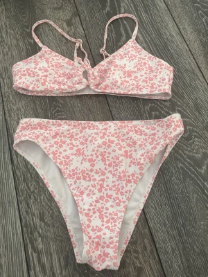 Blommig bikini i rosa och vitt - Söt bikini med blommigt mönster i rosa och vitt. Överdelen har smala axelband och en liten knutdetalj framtill. Underdelen är klassisk med högre midja. Perfekt för strandhäng eller poolen!