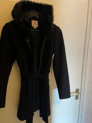Svart kappa med fuskpäls från Anna Field - Snygg svart kappa från Anna Field med huva kantad av mjuk fuskpäls. Jackan har dragkedja och bälte i midjan för en clean siluett. Långärmad och perfekt för löp kyliga dagar. Passar dig som vill ha en klassisk men ändå trendig look.