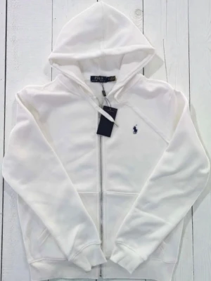 Polo Ralph Lauren hoodie  - Stilren vit hoodie från Polo Ralph Lauren i klassisk design. Tillverkad i mjukt och bekvämt material som passar perfekt till både vardag och mer uppklädda outfits. Den ikoniska brodyren på bröstet ger en tidlös och exklusiv känsla.  🔹 Märke: Polo Ralph Lauren 🔹 Färg: Vit 🔹 Skick: Nyskick / mycket fint skick 🔹 Passform: Klassisk 🔹 Äkta vara  📦 Snabb leverans – skickas samma dag vid beställning innan utsatt tid.
