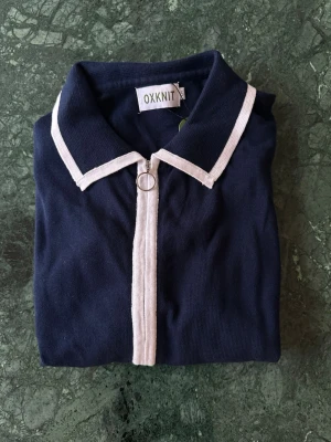 Mörkblå kofta med vita detaljer OXKNIT - Snygg mörkblå kofta från OXKNIT med vita kontrastdetaljer längs krage, dragkedja och ärmslut. Klassisk design med lång ärm och ringformad dragkedja framtill. Perfekt för dig som gillar stilrena plagg med en twist.