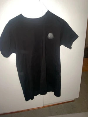 Svart Moncler t-shirt i bomull - Säljer en svart t-shirt från Moncler med klassisk rund hals och diskret Moncler-logga på bröstet. T-shirten är gjord i mjuk bomull och har en clean, stilren look som passar till allt. Perfekt för dig som gillar exklusiva basics.