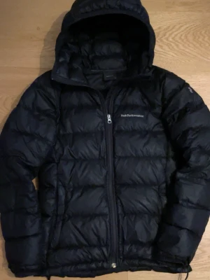 Svart dunjacka Peak Performance XS - Säljer en svart dunjacka från Peak Performance i storlek XS. Jackan har huva, dragkedja framtill och diskret logga på bröstet samt ärmen. Perfekt för kalla dagar och riktigt skön att bära. Klassisk pufferstil som alltid funkar.