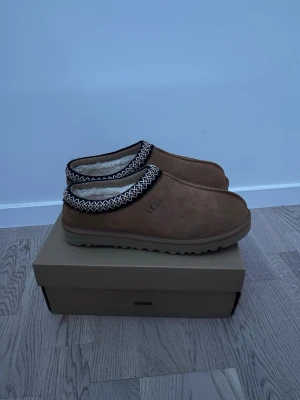 UGG bruna slip-on boots med mönster - Säljer ett par bruna UGG slip-on boots med fluffigt vitt foder och snyggt zigzag-mönster runt öppningen. Skorna har en rund tå och platt sula med grovt mönster undertill. Yttermaterialet är mocka och insidan är fodrad med syntetpäls för extra värme.