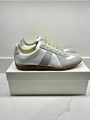 Vita och grå sneakers med mockadetaljer - Snygga sneakers i vitt och ljusgrått med klassisk design. Skorna har paneler i mocka och skinn, snörning framtill och en gummisula i brunt. Perfekta för dig som gillar stilrena och tidlösa sneakers med en clean look.