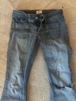 Low waist only jeans  - Säljer ett par blå bootcut jeans från ONLY med slitning och hål på ena benet. Jeansen har två stora fickor fram och klassisk knappstängning. Perfekta för dig som gillar en avslappnad och trendig look. Står 27/30 men passar en med w26-25