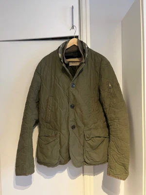 Olivgrön quiltad jacka Burberry Brit L - Säljer en olivgrön quiltad jacka från Burberry Brit i storlek L. Jackan har stora fickor med knappar, ribbad krage och stängs med både dragkedja och knappar. Tillverkad i 100% polyester, perfekt för höst och vinter. Snygg och tidlös design. OBS den sitter som en liten medium inte som en large. Har tappat färgen lite men ändå i bra skick. 