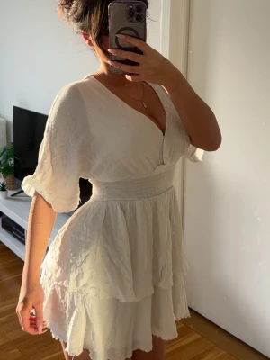 Vit omlottklänning med volanger - Superfin vit kortklänning med omlott och v-ringning. Klänningen har vida korta ärmar, markerad midja och flera lager volanger i kjolen. Perfekt för sommarens alla tillfällen och ger en luftig och romantisk känsla.