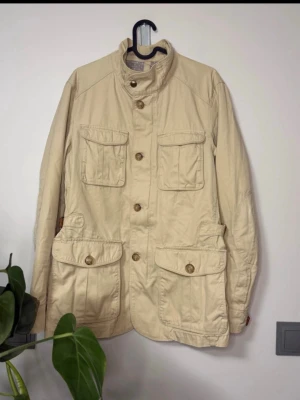 Beige Field jacket från Massimo Dutti - Snygg beige parkajacka från Massimo Dutti med fyra stora fickor framtill och hög krage. Jackan har knappar hela vägen och justerbara detaljer i midjan. Perfekt för dig som gillar stilren och funktionell design.