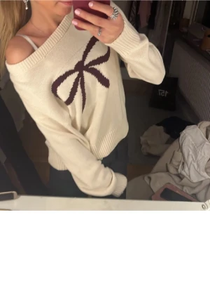 Beige stickad tröja med rosett - Superfin beige stickad tröja med stor mörklila rosett framtill. Tröjan har offshoulder-passform och ribbade muddar vid ärmslut och nederkant. Perfekt för dig som gillar söta detaljer och vill ha en mysig men trendig look. Tröjan är från Gina Young mer storlek 146/152 vilket motsvarar XS💞